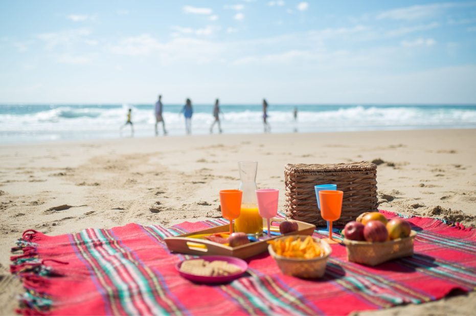 Beach Picnic with Picnic Rug, Glasses and Fruit in the Foreground, Family Group in Distance on Shoreline